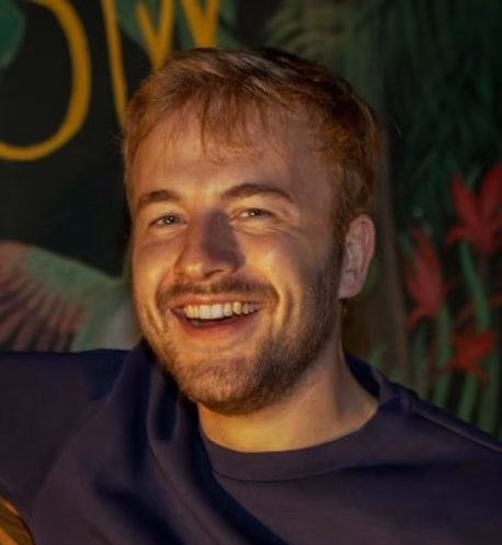 Ein lächelnder Mann mit Bart, der ein dunkelblaues T-Shirt trägt, vor einem grünen Hintergrund mit Blumen.