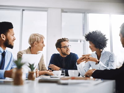 Several people are sitting at a table in an office room, drinking coffee and discussing