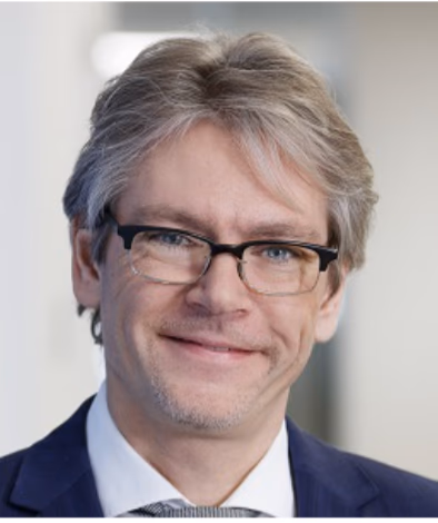 Mann mit grauen Haaren, Brille und Anzug lächelt in die Kamera.