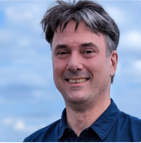 Ein lächelnder Mann mit grauem Haar und blauem Hemd vor blauem Himmel.