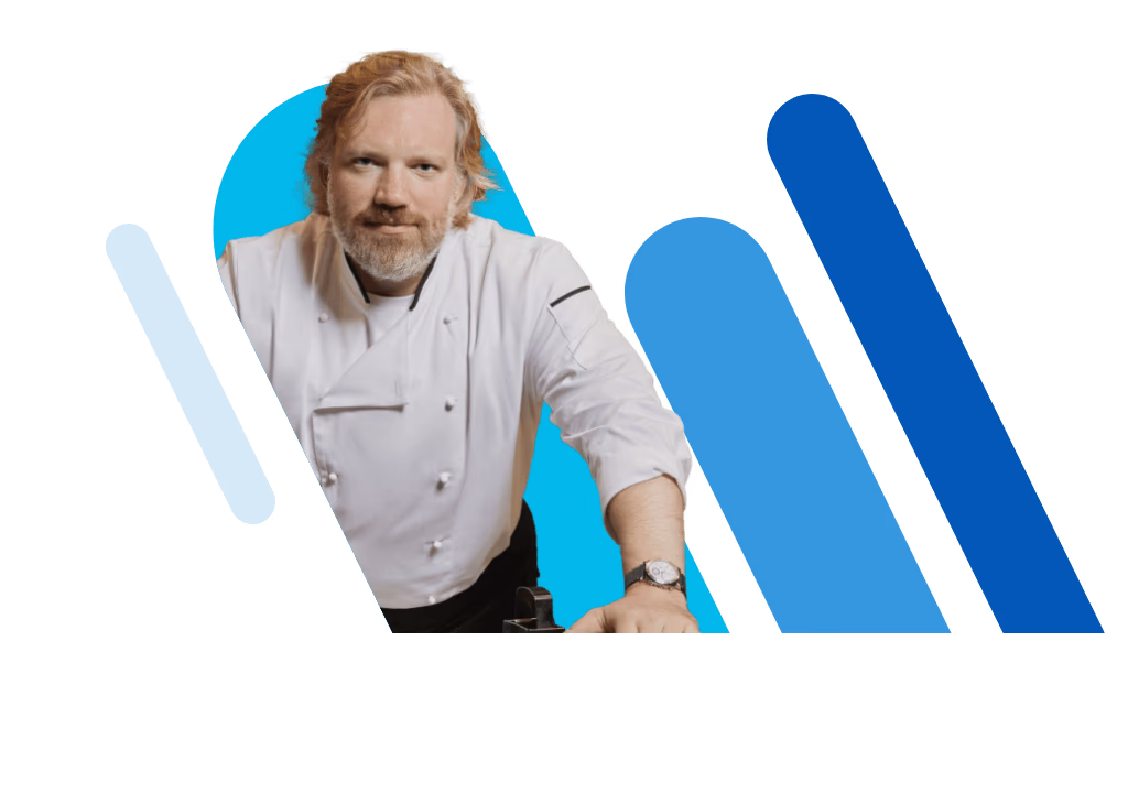 Un chef cuisinier en uniforme blanc avec un tablier noir, portant une montre, sourit et fait un signe de la main.