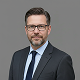 Porträt eines Mannes mit Brille, Anzug und Krawatte vor grauem Hintergrund.