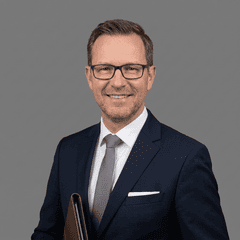 Mann im Anzug mit Krawatte und Brille lächelt in die Kamera.