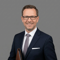 Ein Mann im Anzug mit Krawatte und Brille hält einen Ordner in der Hand.