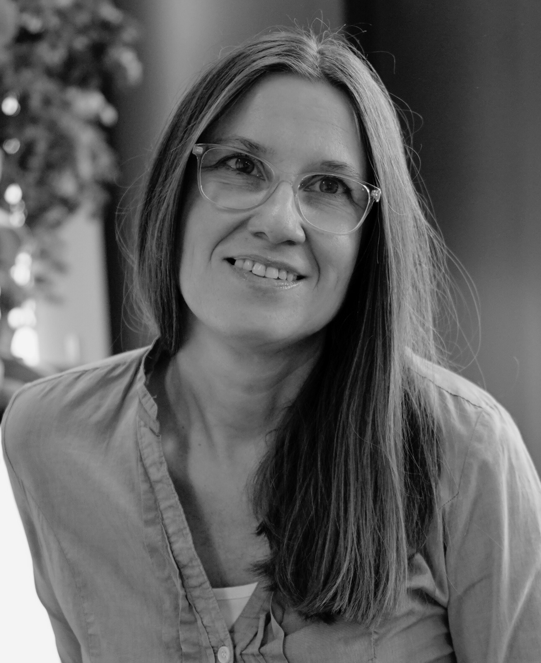 Eine lächelnde Frau mit Brille und langem Haar, die eine Bluse trägt.