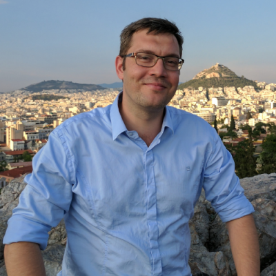 Mann mit Brille und blauem Hemd vor einer Stadt mit einem Hügel im Hintergrund.